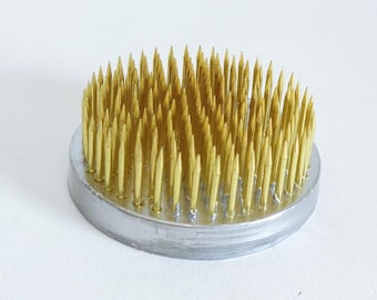 May include: A round, silver metal flower frog with numerous gold-colored pins. The pins are closely spaced and arranged in a dense pattern, creating a textured surface. This floral arranging tool is designed to hold flower stems in place.