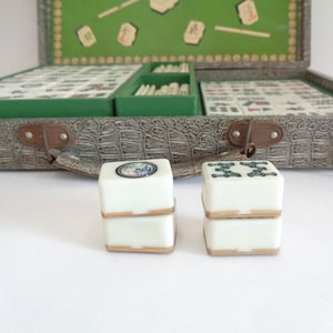 May include: An open vintage Mahjong set with a faux-leather case and green felt interior. Two white dice with decorative tops are in the foreground. The case is gray with brown clasps and a green interior.