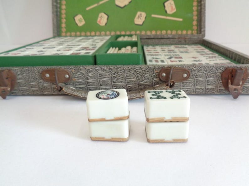 May include: An open vintage Mahjong set with a faux-leather case and green felt interior. Two white dice with decorative tops are in the foreground. The case is gray with brown clasps and a green interior.