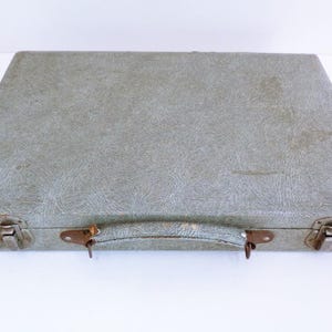 May include: A vintage, rectangular suitcase in a muted green colour. The case features a textured surface, metal latches, and a handle. The corners show signs of wear, suggesting its age. The suitcase is closed and ready for travel.