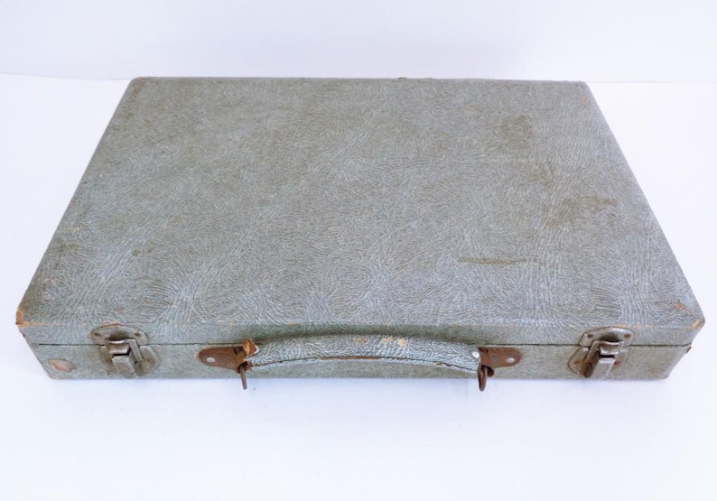 May include: A vintage, rectangular suitcase in a muted green colour. The case features a textured surface, metal latches, and a handle. The corners show signs of wear, suggesting its age. The suitcase is closed and ready for travel.