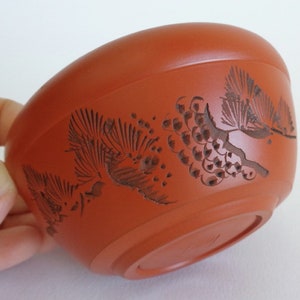 Pode incluir: Uma pequena chávena de chá de cor terracota com um desenho talhado de ramos e pinhas de pinheiro. A chávena está ligeiramente inclinada, mostrando o fundo.