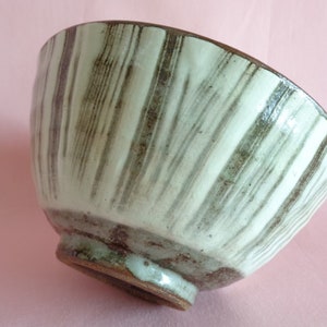 May include: A ceramic bowl with a white glaze and vertical brown brushstrokes. The bowl has a flared rim and a footed base. The interior is smooth, while the exterior has a textured, ribbed appearance. The bowl is suitable for serving food.
