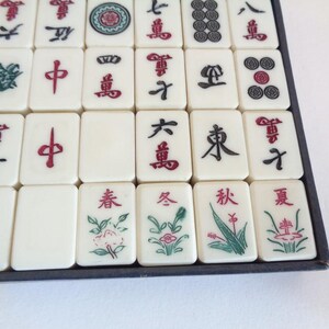 May include: A close-up of a Mahjong set with ivory-colored tiles. The tiles feature red, green, and black symbols and characters. Some tiles have floral designs. The set is arranged in a dark blue tray.