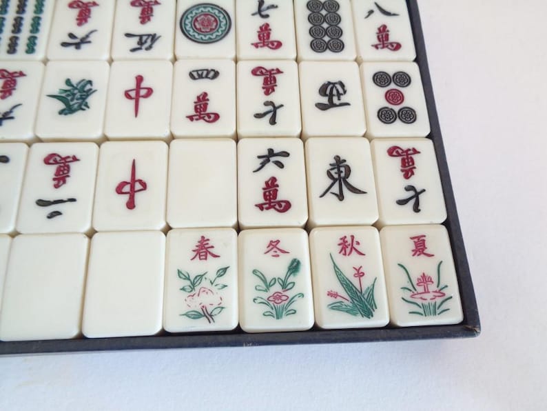 May include: A close-up of a Mahjong set with ivory-colored tiles. The tiles feature red, green, and black symbols and characters. Some tiles have floral designs. The set is arranged in a dark blue tray.