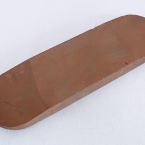Könnte beinhalten: Ein brauner Schleifstein mit abgerundeten Enden. Der Stein ist rechteckig mit einer leicht gewölbten Oberseite. Die Farbe ist ein mittleres Braun, und der Stein scheint aus einem dichten Material zu bestehen.