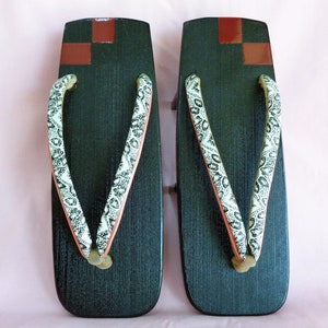 Könnte beinhalten: Paar traditionelle japanische Geta-Sandalen. Die dunkelgrünen Holzplattformen haben eine rechteckige Form mit einer leichten Krümmung. Die Riemen sind weiß mit einem schwarzen Blumenmuster. Die Oberseite jeder Sandale hat ein rot-braunes Karomuster.