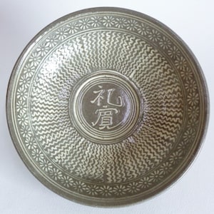 May include: A round, gray ceramic bowl with a white floral pattern around the rim. The bowl has a wavy pattern in the center and a circular design with the Japanese characters "九原" in the middle.