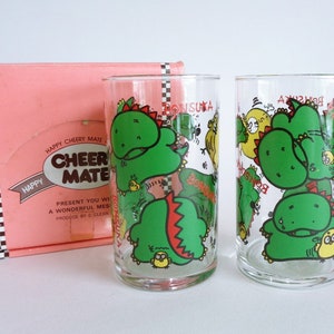 May include: Two clear glass tumblers with cartoon dinosaur graphics in green, red, and yellow. The glasses are next to a pink box with the words "Happy Cheery Mate" printed on it.