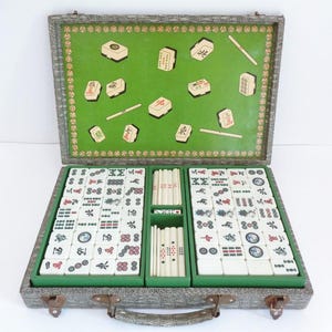 May include: Open mahjong set in a gray case with a green interior. The set includes ivory-colored tiles with red, black, and blue symbols, along with green tile holders and dice. The case is open, revealing the game pieces.