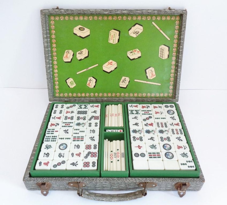 May include: Open mahjong set in a gray case with a green interior. The set includes ivory-colored tiles with red, black, and blue symbols, along with green tile holders and dice. The case is open, revealing the game pieces.