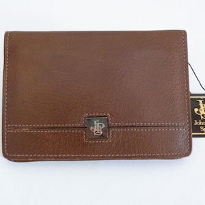 May include: A brown leather wallet with a rectangular shape. The wallet has a stitched border and a silver-colored metal emblem on the front. A black tag with the words "John Player Special" is attached.