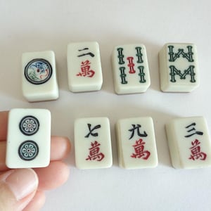 May include: A collection of off-white Mahjong tiles with black, red, and green markings. The tiles feature various symbols and Chinese characters. One tile is held by a hand.