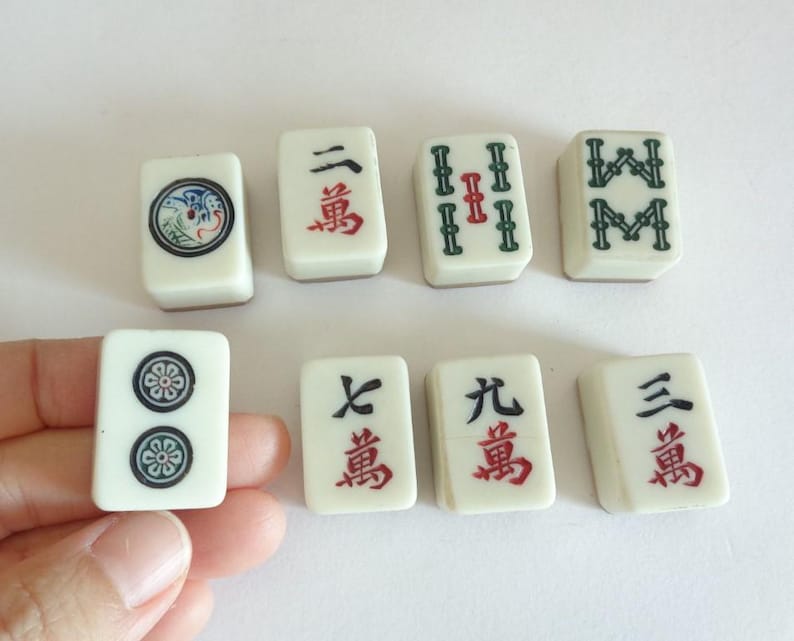 May include: A collection of off-white Mahjong tiles with black, red, and green markings. The tiles feature various symbols and Chinese characters. One tile is held by a hand.