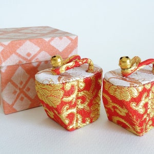 May include: Two small red and gold fabric boxes with gold bells and handles. The boxes are sitting on a white surface next to a pink and white patterned box.