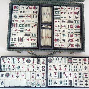 May include: A vintage Mahjong set in a green case, open to reveal tiles with red, green, and black symbols. The set includes dice and tile racks. The tiles are white with various symbols and characters.
