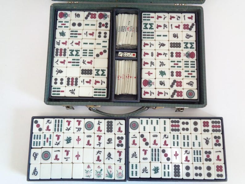 May include: A vintage Mahjong set in a green case, open to reveal tiles with red, green, and black symbols. The set includes dice and tile racks. The tiles are white with various symbols and characters.