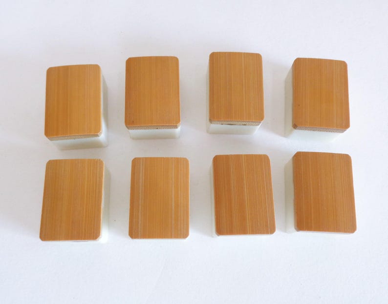 May include: Eight rectangular mahjong tiles. Each tile has a light brown, bamboo-like top and a white base. The tiles are arranged in two rows of four, with the tops facing upwards. The bamboo grain is visible on each tile.