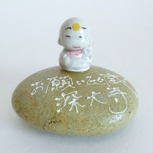 May include: A small, white ceramic figurine of a smiling, cartoon-like figure with a yellow dot on its forehead, sitting atop a green, oval-shaped stone with white calligraphy. The calligraphy reads "五福臨門" and "招財進寶".