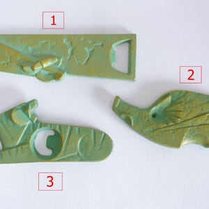 May include: Three green metal bottle openers with different designs. The first opener is a rectangular shape with a leaf design. The second opener is shaped like a fish with a leaf design. The third opener is a long, thin shape with a leaf design.