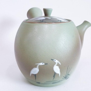 May include: A round, sage green ceramic teapot with a clear lid and a small handle. The teapot features a painted design of two white birds with long necks and black legs, standing near a body of water.