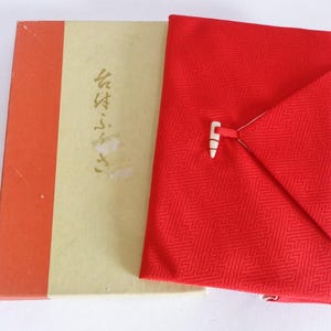 以下が含まれることがあります： 赤い背表紙と日本語の文字が書かれたクリーム色の長方形の箱と、幾何学模様が施された鮮やかな赤色の折り畳まれた布の正方形。布には小さな白いトグル留め具が付いており、繰り返される角張ったデザインのテクスチャーが特徴です。