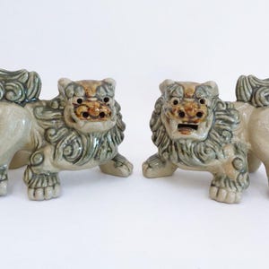 May include: A pair of ceramic foo dog figurines featuring a glossy glaze in shades of sage green, tan, and brown. Each mythical lion creature is depicted in a standing position with detailed manes, curled tails, and open mouths showing teeth.