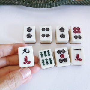 May include: A collection of white Mahjong tiles with black, red, and green symbols. One tile is held in a hand. The tiles are arranged on a white surface, with a green patterned case in the background.