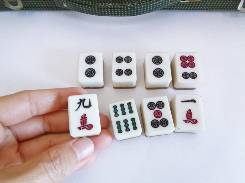 May include: A collection of white Mahjong tiles with black, red, and green symbols. One tile is held in a hand. The tiles are arranged on a white surface, with a green patterned case in the background.