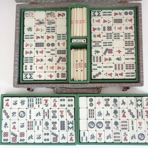 May include: A vintage Mahjong set with ivory-colored tiles featuring red and black symbols and characters. The set includes dice and is housed in a decorative, silver-toned case with green compartments. The tiles are arranged in a neat, organized manner.