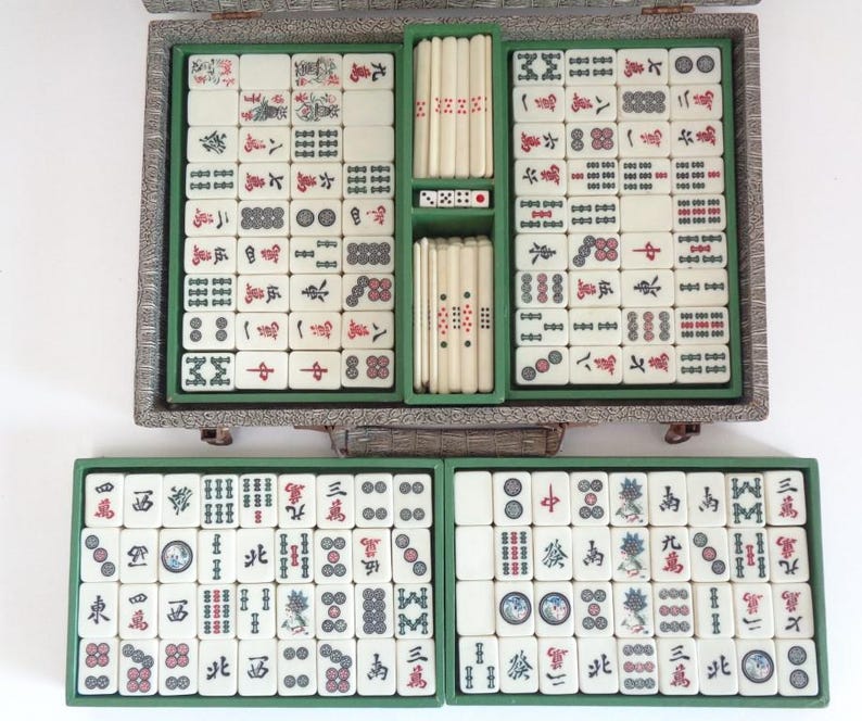 May include: A vintage Mahjong set with ivory-colored tiles featuring red and black symbols and characters. The set includes dice and is housed in a decorative, silver-toned case with green compartments. The tiles are arranged in a neat, organized manner.