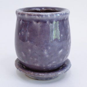 Puede incluir: Un pequeño macetero de cerámica morado con un platillo a juego. El macetero tiene un esmalte moteado y una forma redondeada.