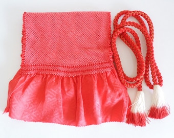 May include: A red fabric bag with a ruffled bottom and a matching red cord with white tassels. The bag has a textured surface and a woven design.