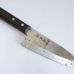 Puede incluir: Un cuchillo de cocina de estilo japonés con un mango de madera marrón oscuro asegurado por dos remaches. La hoja es plateada con desgaste visible y presenta caracteres japoneses. El cuchillo está diseñado para cortar con precisión.