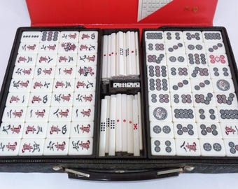 May include: A black carrying case with a brown leather exterior and a handle. The case is open, revealing a set of white mahjong tiles with red and black markings. The tiles are arranged in rows and columns, and there are also a set of white dice and a set of white bamboo sticks.