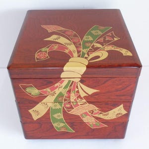May include: A three-tiered, reddish-brown wooden bento box. The box is decorated with a gold-colored knot and ribbons in red, green, and gold with floral patterns. The box has a rectangular shape with angled sides.