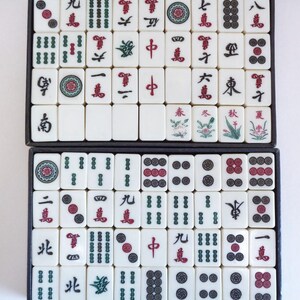 May include: A complete set of ivory-colored Mahjong tiles arranged in two black trays. The tiles feature Chinese characters and symbols in red and green, including dots, bamboo, and dragons. The game set is ready for play.
