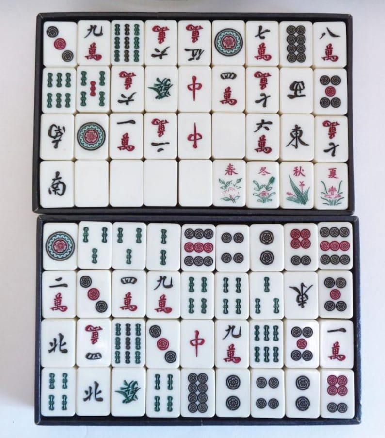 May include: A complete set of ivory-colored Mahjong tiles arranged in two black trays. The tiles feature Chinese characters and symbols in red and green, including dots, bamboo, and dragons. The game set is ready for play.