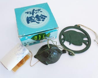 May include: A dark green cast iron fish-shaped wind chime with a matching circular frame featuring a fish cutout, connected by a string to a white paper wind catcher. The set includes a light blue cardboard box with Japanese text and fish graphics.