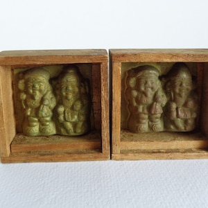 May include: Two wooden boxes each containing two small green ceramic figurines. The figurines are of people with hats and beards.