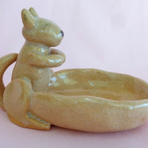 May include: A ceramic squirrel-shaped dish in a light tan color. The squirrel is sitting upright with its paws together, and the dish is an oval shape. The dish has a glossy finish and is on a pink background.