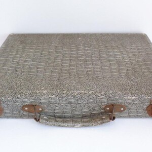 May include: A vintage gray suitcase with a textured, crocodile-like pattern. The suitcase has a handle and two latches with a rusty appearance. The corners of the suitcase show signs of wear. The suitcase is closed and sits on a white surface.