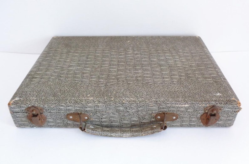May include: A vintage gray suitcase with a textured, crocodile-like pattern. The suitcase has a handle and two latches with a rusty appearance. The corners of the suitcase show signs of wear. The suitcase is closed and sits on a white surface.
