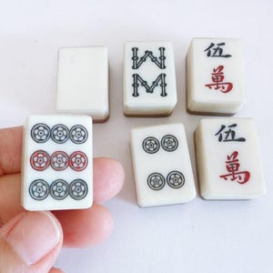 May include: A selection of vintage Mahjong tiles. The tiles are white with black, red, and green symbols. Some tiles feature Chinese characters. The tiles are rectangular and have a glossy finish.