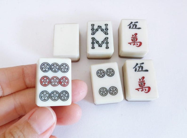 May include: A selection of vintage Mahjong tiles. The tiles are white with black, red, and green symbols. Some tiles feature Chinese characters. The tiles are rectangular and have a glossy finish.
