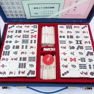Puede incluir: Estuche azul abierto que contiene un juego de Mahjong completo. El juego incluye fichas blancas con símbolos negros y rojos, dados y una pieza circular roja. El estuche está forrado con tela roja e incluye documentos.