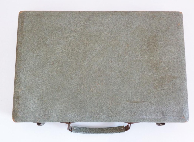May include: A vintage, rectangular briefcase in a muted green hue. The case features a textured surface and a handle with metal hardware. The briefcase is closed and appears to be empty.
