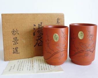 Könnte beinhalten: Zwei rote Keramik-Teetassen mit Vogel- und Blumendesign. Die Tassen befinden sich in einer Holzkiste mit japanischen Schriftzeichen.