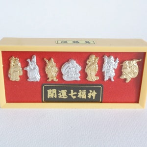May include: A display of seven Japanese lucky gods in a rectangular box. The figures are in gold and silver tones, set against a red background. The box has a cream-coloured frame and a black label with Japanese text.