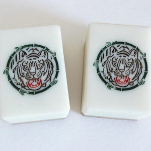 May include: Two white Mahjong tiles with a tiger face design. The tiger is encircled by a green bamboo-like border. The tiger's face is black and white with a red mouth.